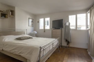 Chambre lumineuse d’un duplex rue d’Aboukir, avec parquet ancien, lit en métal, commode, télévision et fenêtres donnant sur les toits parisiens.