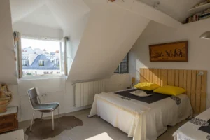 Chambre sous combles d’un duplex rue d’Aboukir, avec tête de lit en bois, lumière naturelle, fenêtre ouverte et vue sur les toits parisiens.
