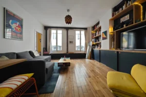 Séjour lumineux d’un appartement 3 pièces à vendre à Paris 18 Place de Clichy, parquet ancien, double exposition, bibliothèque sur mesure, cuisine semi-ouverte et fenêtres sur rue et cour dans un immeuble en pierre de Paris.