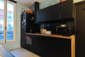 Cuisine équipée avec nombreux rangements dans un appartement à vendre à Paris 18 Place de Clichy, meubles noirs contemporains, plan de travail en bois, crédence sombre, électroménager intégré et fenêtre apportant une lumière naturelle.