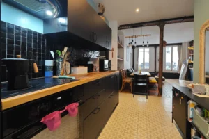 Perspective depuis la cuisine ouverte vers le séjour d’un appartement à vendre à Paris 18 Place de Clichy, plan de travail en bois, crédence noire, rangements fonctionnels, piliers en fonte et espace de vie lumineux dans un immeuble ancien rénové.