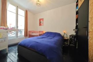 Chambre lumineuse d’un appartement à vendre à Paris 18 Place de Clichy avec parquet ancien peint, lit double, fenêtre sur rue apportant une belle lumière naturelle, espace nuit confortable et rangements à proximité.