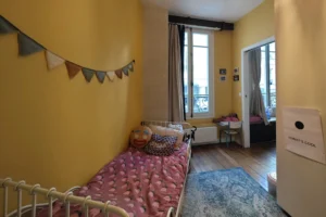 Chambre d’enfant lumineuse d’un appartement à vendre à Paris 18 Place de Clichy, avec parquet ancien, fenêtre sur rue, coin nuit cosy, murs colorés et accès vers la seconde chambre par une porte communicante.