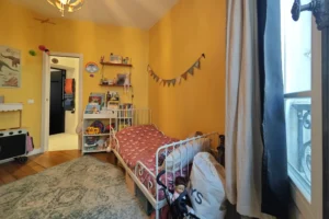 Seconde chambre d’enfant d’un appartement à vendre à Paris 18 Place de Clichy, avec parquet ancien, fenêtre sur rue, espace nuit chaleureux, étagères murales et aménagement adapté à une chambre familiale.