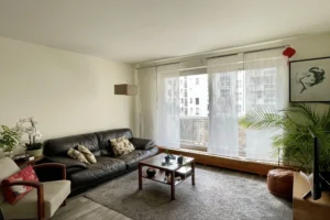Vue d’ensemble du séjour lumineux d’un appartement à vendre à Paris 12 Picpus, canapé en cuir, grande baie vitrée avec rideaux voilages et espace salon chaleureux.