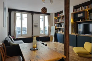 Vue d’ensemble du séjour double exposition d’un appartement à vendre à Paris 18 Place de Clichy, parquet ancien, poutres et piliers en fonte apparents, grande bibliothèque sur mesure, coin salon et espace repas dans un immeuble ancien.