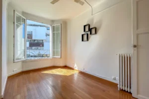 Séjour avec parquet en bois, murs blancs, fenêtre à battants ouverte laissant entrer la lumière, radiateur ancien et étagères murales, dans un appartement à Paris 12 Reuilly-Diderot.