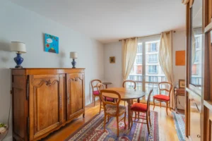 Séjour avec espace salle à manger d’un appartement à vendre à Paris 12 Picpus, parquet en bois, table ronde et chaises, buffet ancien, grande fenêtre et rideaux clairs apportant une belle lumière naturelle.