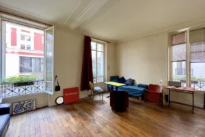 Séjour d’un studio à Paris 10 avec parquet ancien, grandes fenêtres sur cour végétalisée et verrière d’atelier, murs clairs, coin salon et espace nuit minimaliste.