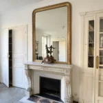 Détail d’une cheminée ancienne en pierre avec grand miroir doré dans un appartement à vendre à Paris 11, boulevard du Temple, parquet en point de Hongrie et portes anciennes.