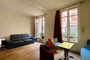 Pièce de vie d’un studio à Paris 10 avec parquet ancien, grande fenêtre donnant sur cour végétalisée, canapé, coin repas et ambiance lumineuse côté rue du Faubourg Saint-Denis.