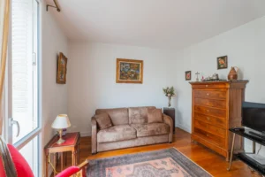 Salon d’un appartement à vendre à Paris 12 Picpus, parquet en bois, canapé deux places, commode ancienne, tapis décoratif, fenêtre apportant une belle luminosité naturelle et vue dégagée sur l’extérieur.