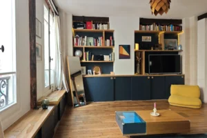 Mur de rangements et bibliothèque sur mesure dans le séjour d’un appartement à vendre à Paris 18 Place de Clichy, avec parquet ancien, assise sous fenêtre, miroir pleine hauteur, espace TV intégré et ambiance chaleureuse dans un immeuble ancien.