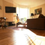 Salon d’un appartement ancien à vendre à Paris 16 Molitor, parquet d’origine, canapé et table basse, grandes fenêtres ouvertes sur balcon avec garde-corps, vue arborée et atmosphère calme.