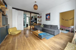 Pièce de vie traversante d’un appartement à vendre à Paris 18 Place de Clichy avec parquet ancien, séjour spacieux, piliers en fonte apparents, coin salle à manger et circulation fluide vers la cuisine et les chambres dans un immeuble ancien rénové.