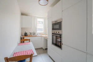 Cuisine indépendante équipée d’un appartement à vendre à Paris 12 Picpus, mobilier blanc contemporain, plan de travail en bois, four encastré, coin repas avec table et fenêtre apportant une lumière naturelle.