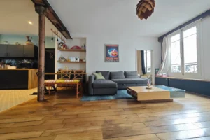 Vue large du salon avec cuisine ouverte d’un appartement à vendre à Paris 18 Place de Clichy, parquet ancien, piliers en fonte, double exposition, coin salle à manger et luminosité naturelle dans un immeuble ancien rénové.