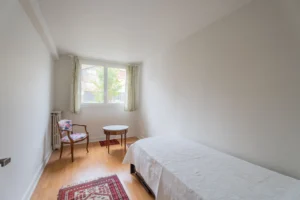 Chambre d’un appartement à vendre à Paris 12 Picpus, parquet en bois, lit double, fauteuil et petite table, fenêtre donnant sur l’extérieur, ambiance calme et lumineuse.