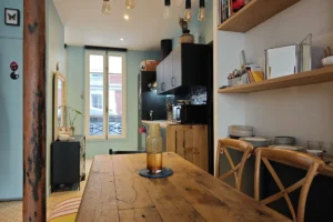 Cuisine ouverte et espace salle à manger d’un appartement à vendre à Paris 18 Place de Clichy, meubles contemporains, étagères murales en bois, plan de travail fonctionnel, suspensions design et belle luminosité naturelle.