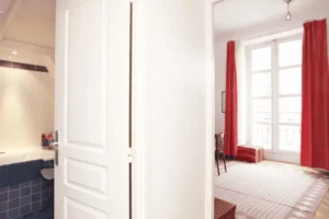 Chambre avec fenêtres et rideaux rouges, pièce claire pouvant servir de chambre ou salle de jeux, vue sur immeubles parisiens.