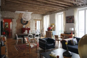 Grande pièce de vie traversante très lumineuse avec salon et salle à manger, multiples fenêtres ouvrant sur balcon, parquet ancien, poutres en bois et murs en pierre, appartement Paris 2 Sentier rue de Tracy.