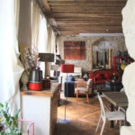 Vue depuis l’entrée vers le séjour avec parquet en point de Hongrie, poutres anciennes au plafond, murs en pierre et lumière naturelle provenant des grandes fenêtres, décoration chaleureuse, appartement Paris 2 Sentier rue de Tracy.