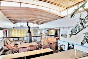 Vue en hauteur sur la grande pièce de vie d’une péniche aménagée en style loft avec plafond voûté en bois, poêle central et espace salle à manger à Levallois-Perret quai Charles Pasqua.