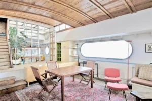 Salle à manger d’une péniche avec plafond voûté en bois, grande fenêtre hublot et table en bois au cœur de l’espace de vie à Levallois-Perret quai Charles Pasqua.