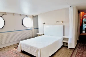 Chambre lumineuse d’une péniche avec lit double, hublot rond et parquet ancien située à Levallois-Perret quai Charles Pasqua.