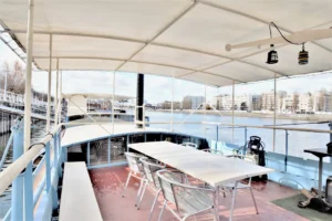 Terrasse couverte d’une péniche d’habitation amarrée quai Charles Pasqua à Levallois-Perret avec table extérieure et vue ouverte sur la Seine et les immeubles des quais.