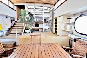 Grand espace de vie intérieur d’une péniche aménagée en style loft avec mezzanine, escalier en bois et verrière centrale à Levallois-Perret quai Charles Pasqua.