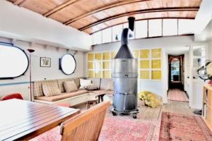 Grande pièce de vie d’une péniche avec plafond voûté en bois, poêle central et banquette intégrée offrant un espace salon convivial à Levallois-Perret quai Charles Pasqua.